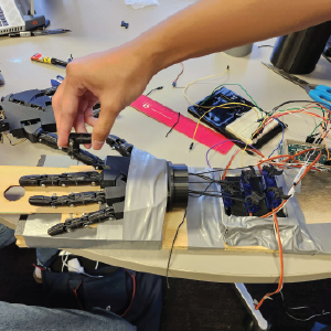 A mechanical hand prototype is mounted on a wooden board, with its fingers controlled by strings connected to multiple servo motors. The setup is secured with duct tape, and various electronic components and wiring are visible, including an Arduino and breadboards. A person is manually adjusting one of the 3D-printed fingers, testing movement or alignment.